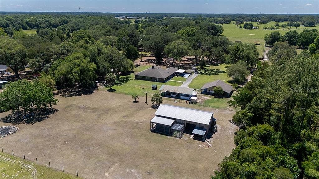 Drone view of the full 5.05-acre compound showing the brick ranch home, two-stall barn, workshop, and pastures laid out beneath the grandfather oaks